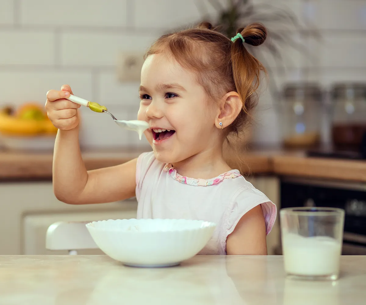 Ein kleines Mädchen sitzt an einem Tisch und isst Brei aus einer Schüssel. Nebn der Schüssel steht ein Glas Milch