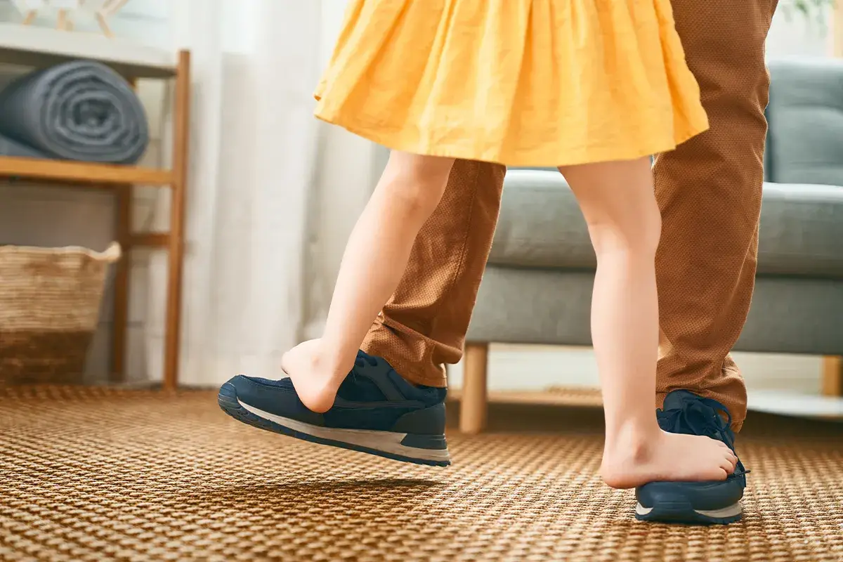 Ausschnitt der Beine eines Kleinkindes, welches steht auf den Füssen einer erwachsenen Person steht und der rechte Fuss dieser Peron ist in der Luft