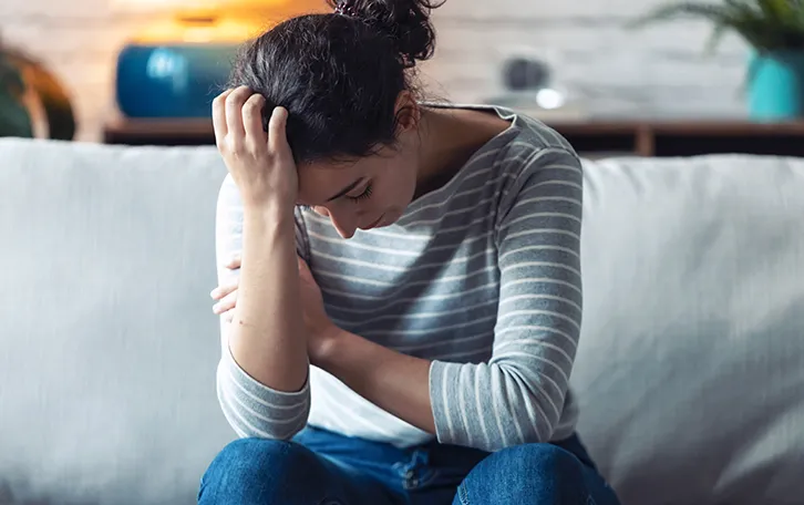 Eine weibliche Person sitzt auf einer Couch und hat den Kopf niedergeschlagen auf die eine Hand gestützt