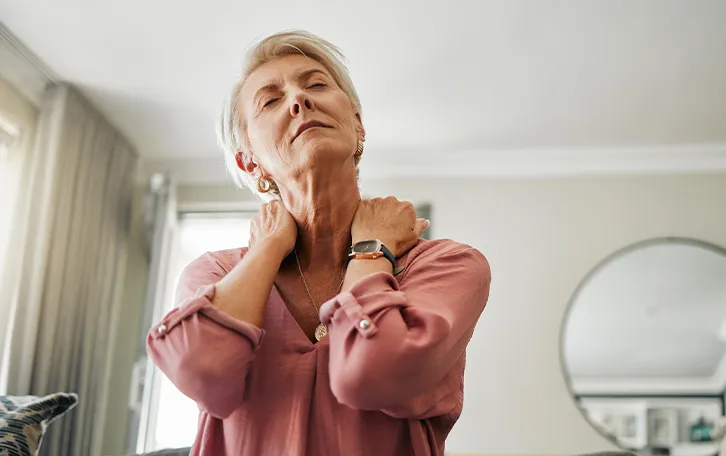Eine ältere weibliche Person mit geschlossenen Augen hält sich mit beiden Händen den Nackenbereich