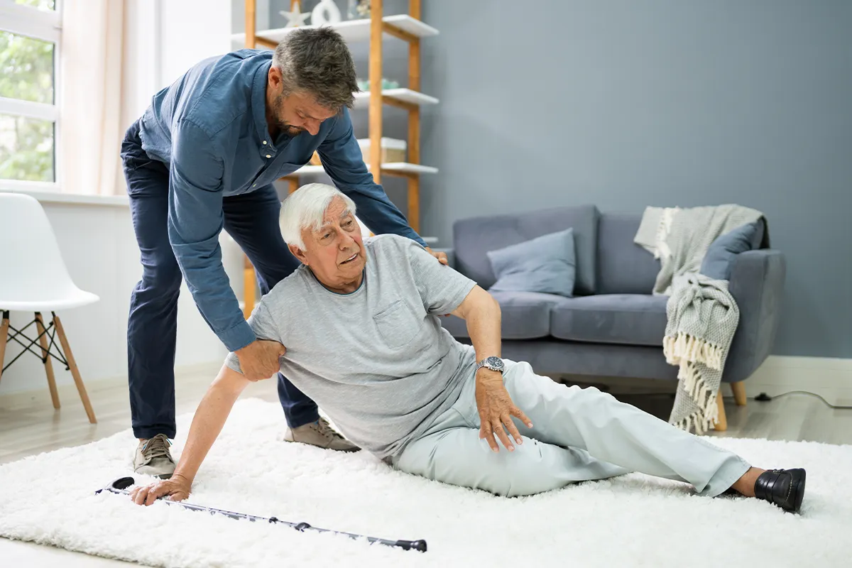 Eine ältere männliche Person liegt auf dem Teppich mit ihrem Stock neben sich und eine jüngere männliche Person scheint ihm auf helfen zu wollen