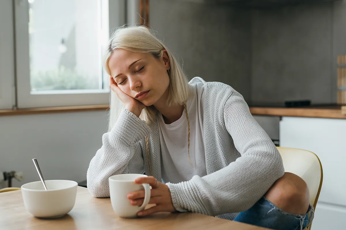 Wann spricht man von Vitamin D Mangel Eine mit geschlossenen Augen weiblich Person sitzt an einem Tisch. In der einen Hand hält sie eine Tasse und auf die andere ist der Kopf gestützt