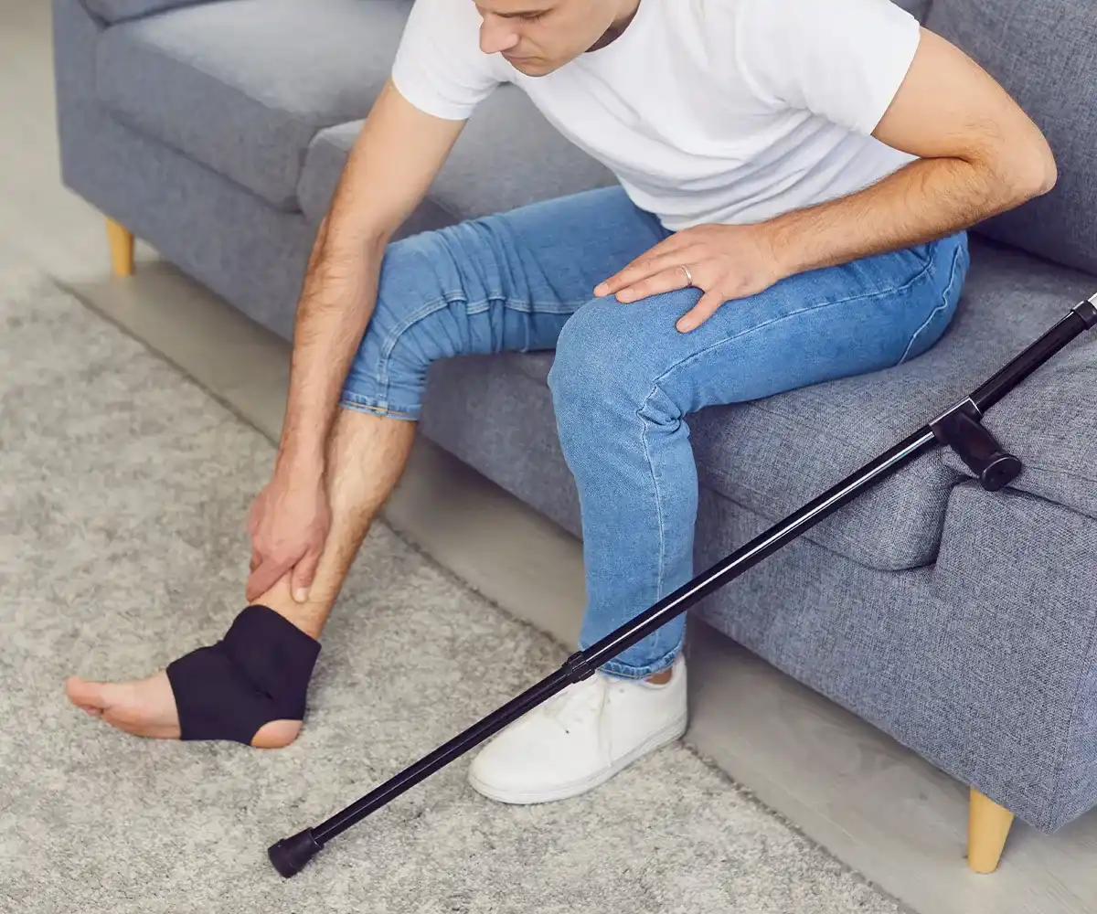 Eine männliche Person mit Bandage an einem Fussgelenk auf einer Couch sitzend und Stock neben sich