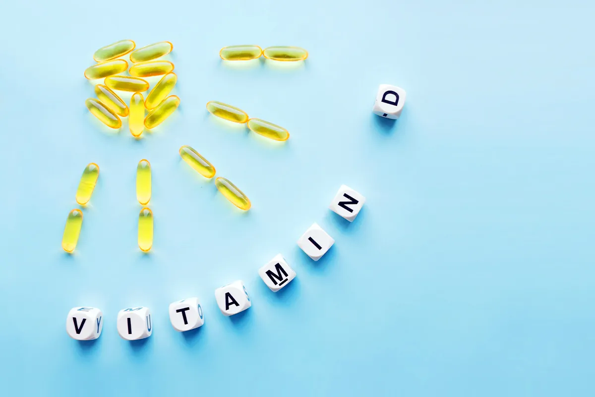 Eine Sonne mit Strahlen gelegt aus gelblichen Pillen und Würfeln mit dem Wort "Vitamin D" im Halbkreis gelegt